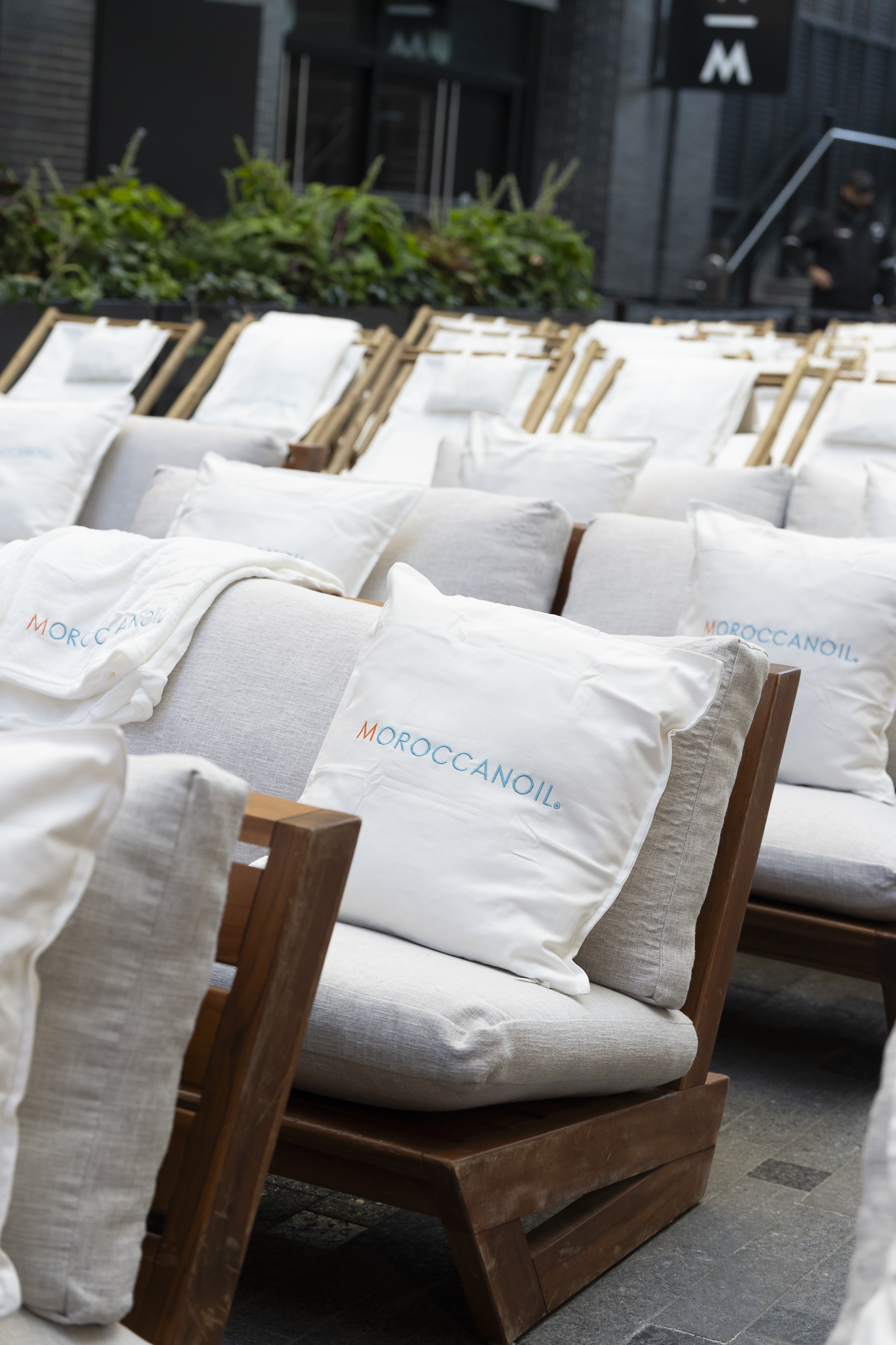 Row of beige outdoor lounge chairs with white cushions and MOROCCANOIL pillows, arranged in an outdoor seating area.