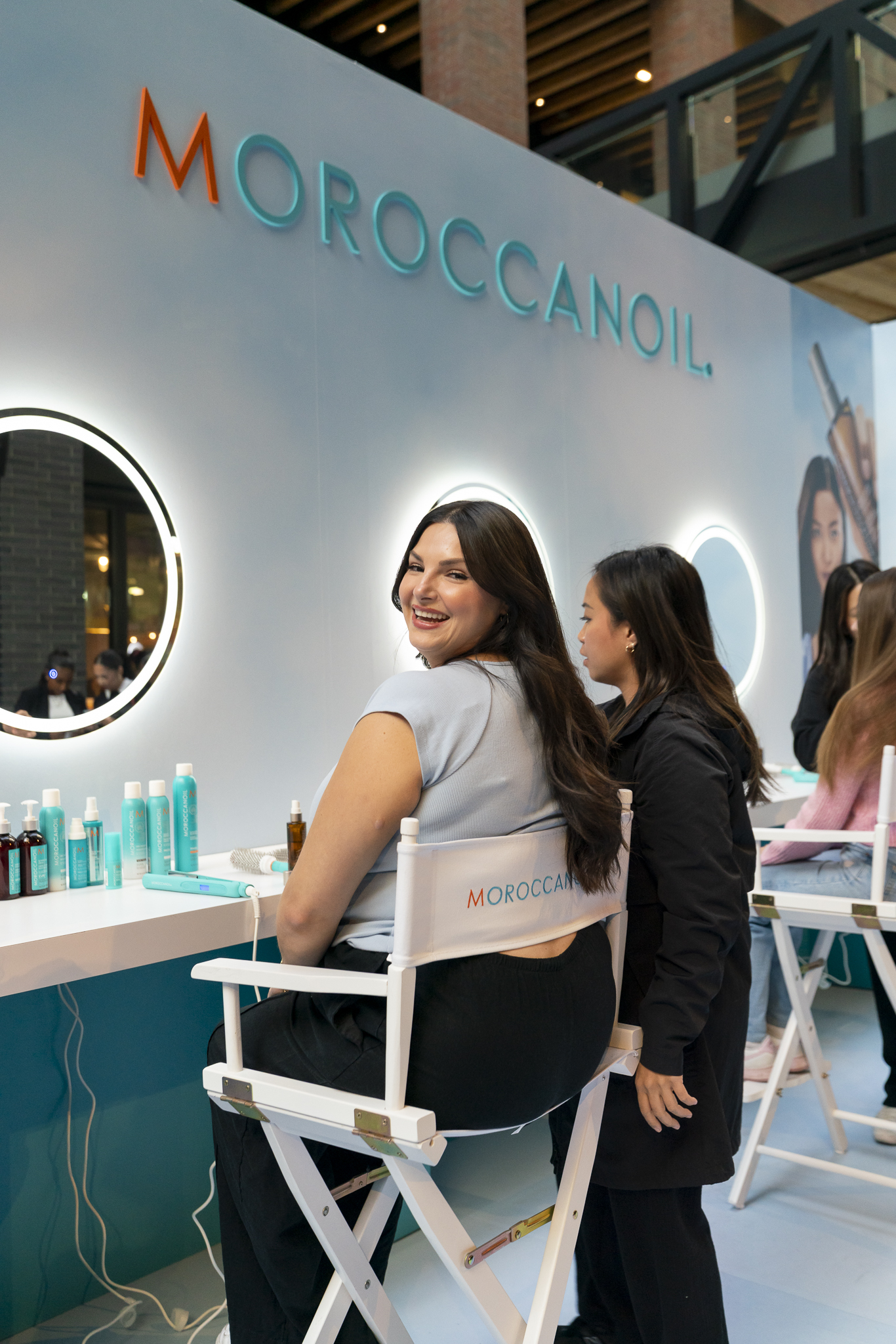 Smiling woman seated at a Moroccanoil booth with turquoise product bottles on the counter and round illuminated mirrors behind her.