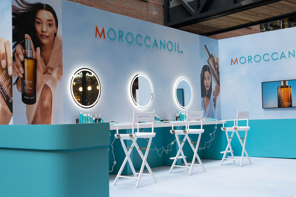 Beauty brand booth with turquoise counter, circular lit mirrors, and white makeup chairs for on-site demos (MOROCCANOIL).