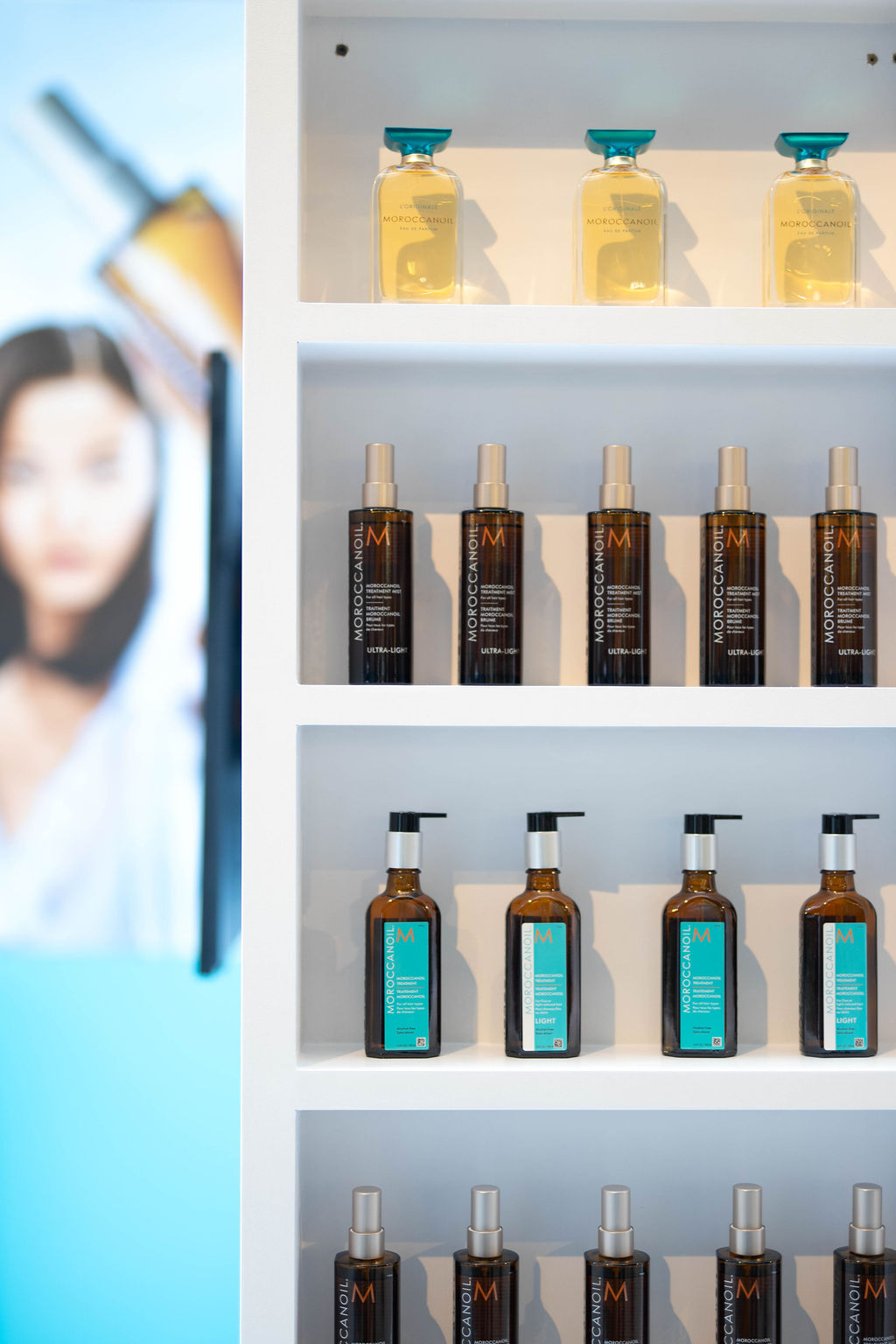 Display shelf with Moroccanoil hair care products: gold oil bottles on the top row and dark brown bottles with silver pumps on the middle row against a white backdrop.