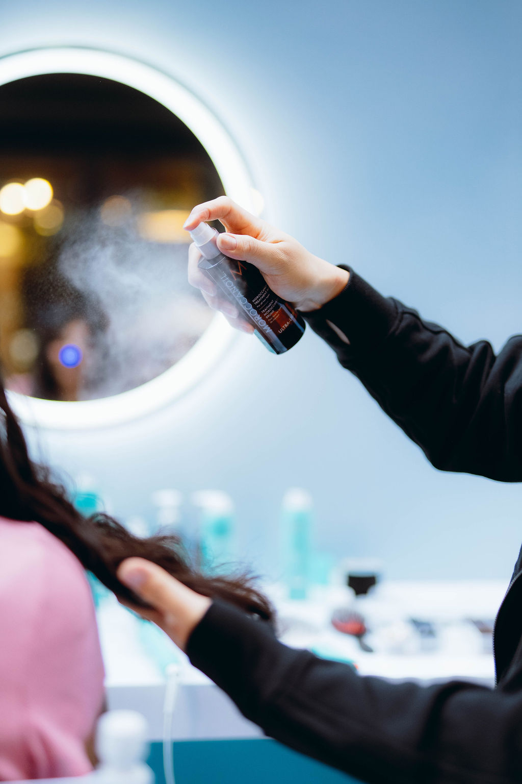 Stylist spritzing hair product onto a client's hair in a bright salon with a ring light Behind them.**