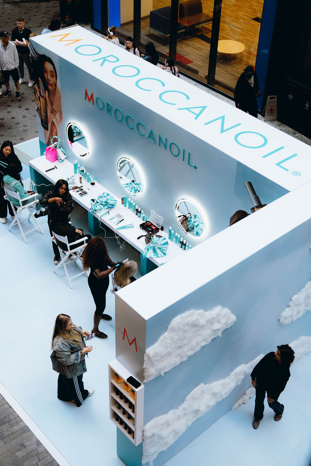 Overhead view of a Moroccanoil beauty booth in a mall, with turquoise branding, makeup stations, round illuminated mirrors, and shoppers browsing and getting services.