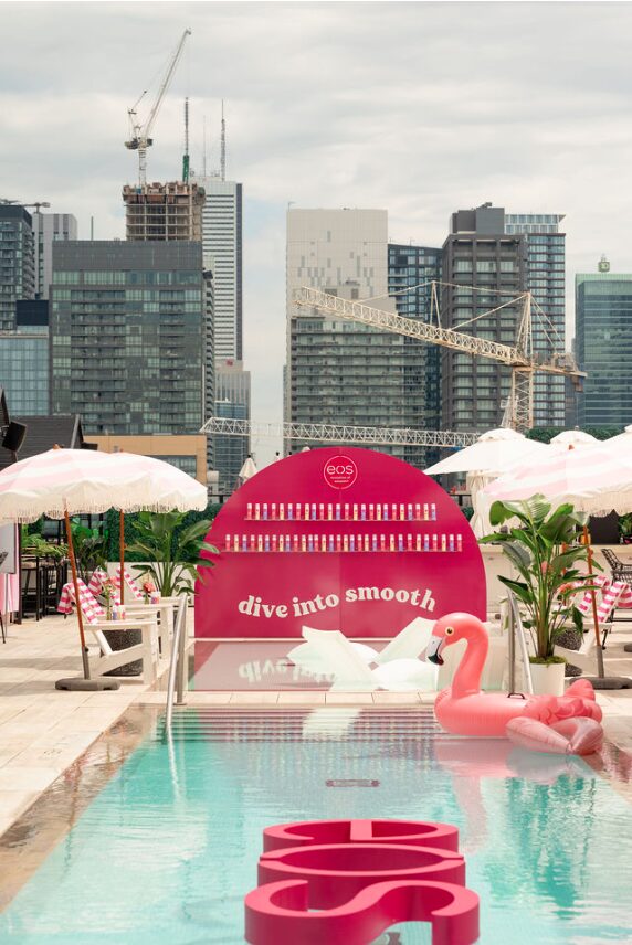 Rooftop pool party with pink signage reading 'dive into smooth', a pink flamingo float in the pool, white umbrellas, and a city skyline with cranes in the background.