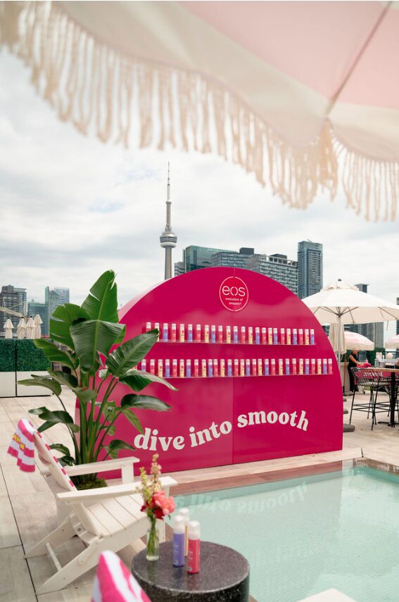 Pink curved cosmetics display with bottles lined on shelves by a rooftop pool; city skyline in the background.