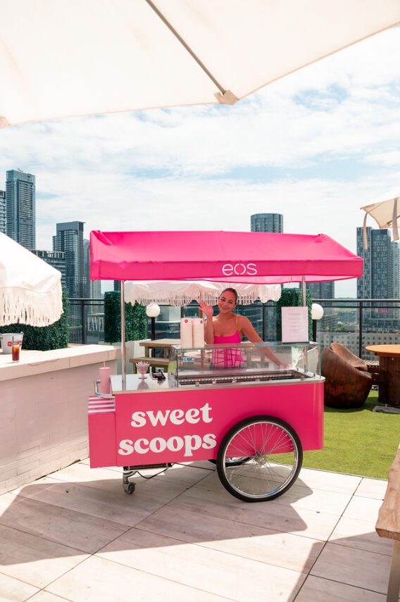 Woman serving pink ice cream cart labeled 'sweet scoops' on a rooftop with city skyline in the background.