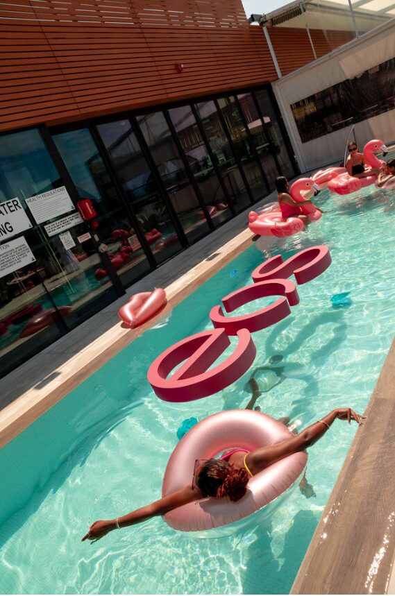 Sunny outdoor pool scene with pink flamingo inflatables and people lounging on floats beside a modern glass-walled building.