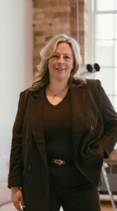 Smiling woman with blonde hair in a brown blazer and dark top, standing in a brick-walled, sunlit room.