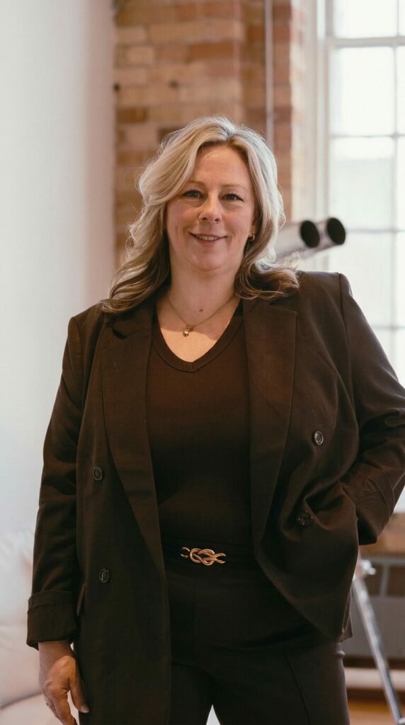 Smiling woman with blonde hair in a brown blazer and dark top, standing in a brick-walled, sunlit room.