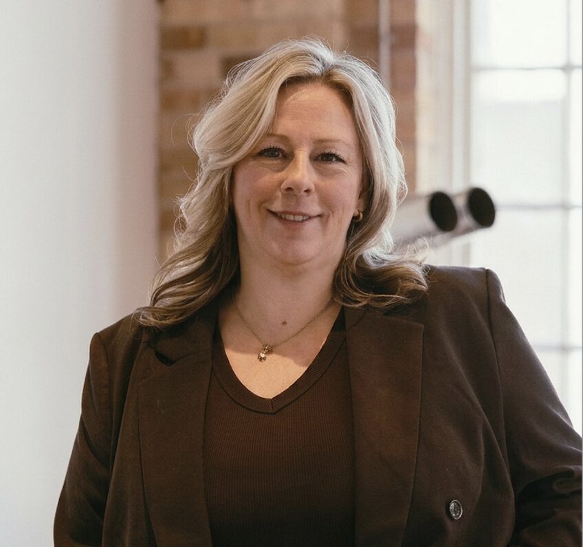 Smiling woman with blonde hair wearing a brown blazer and black top, standing indoors with natural light.