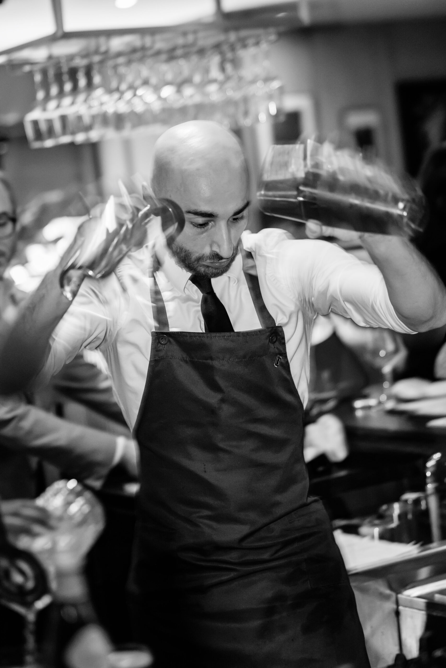 Chef in white shirt and black apron working briskly in a busy kitchen, arms raised as he handles utensils.