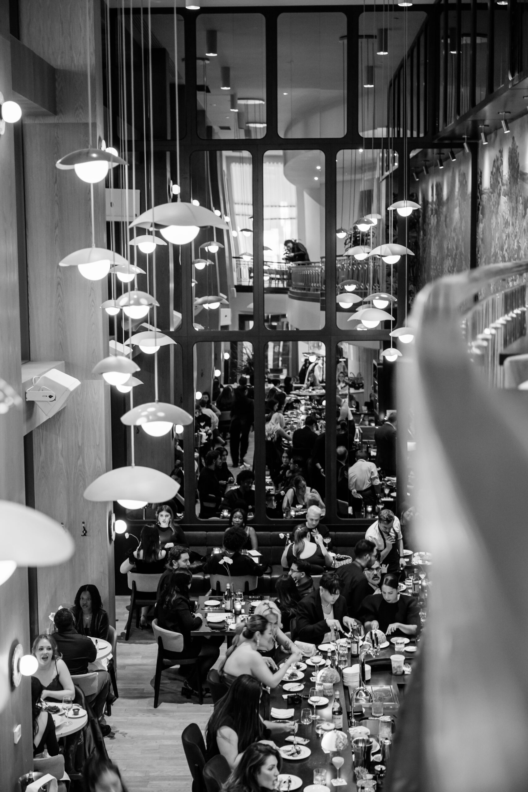Crowded modern restaurant with long tables, diners eating, and hanging dome lamps visible through large windowed dividers in black-and-white.