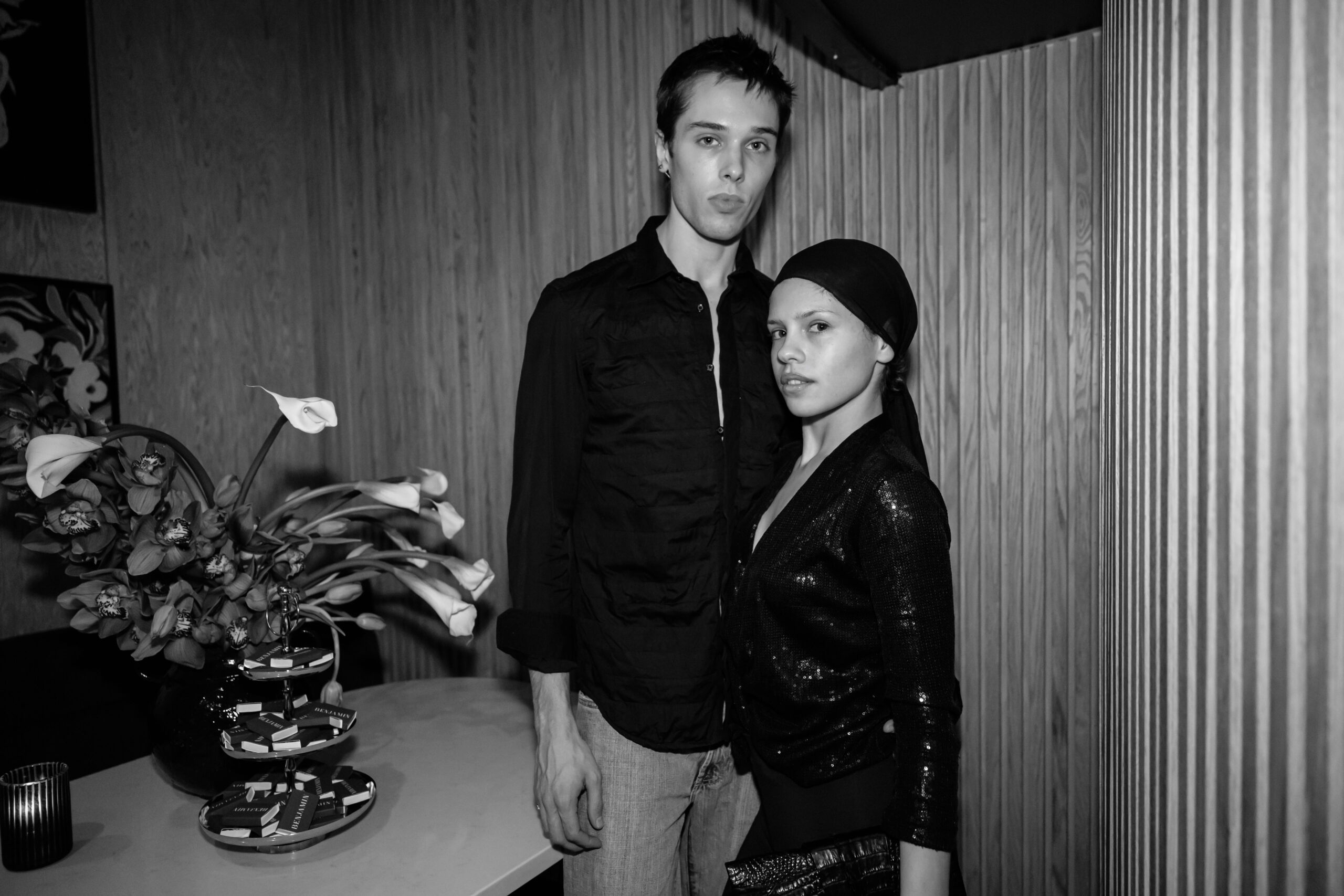 Two models pose indoors beside a table of chocolates and flowers, one tall in black shirt and the other in a sequined outfit with a headscarf.