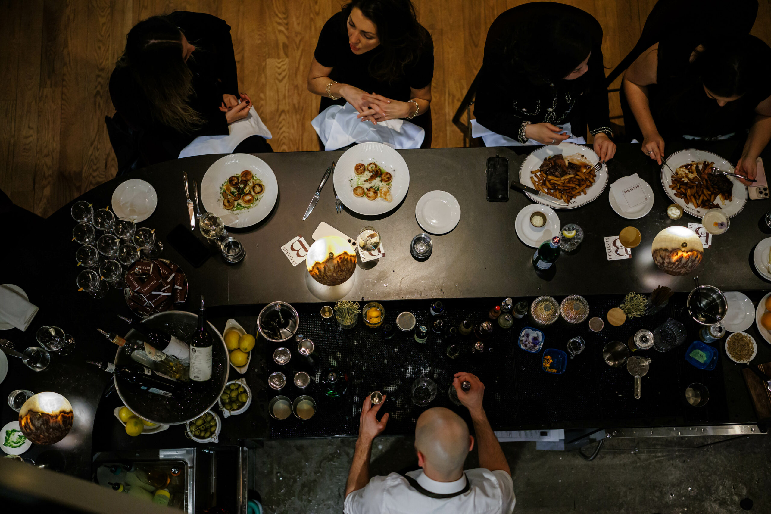Top-down view of people dining at a long bar with plates of food, drinks, and a bartender preparing orders on the counter.