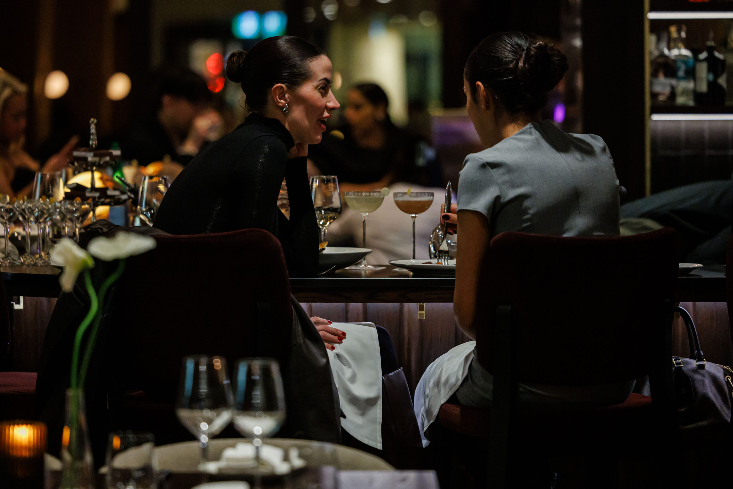 Two women sit across from each other at a dimly lit restaurant table, engaged in conversation with cocktails and wine glasses in front of them.