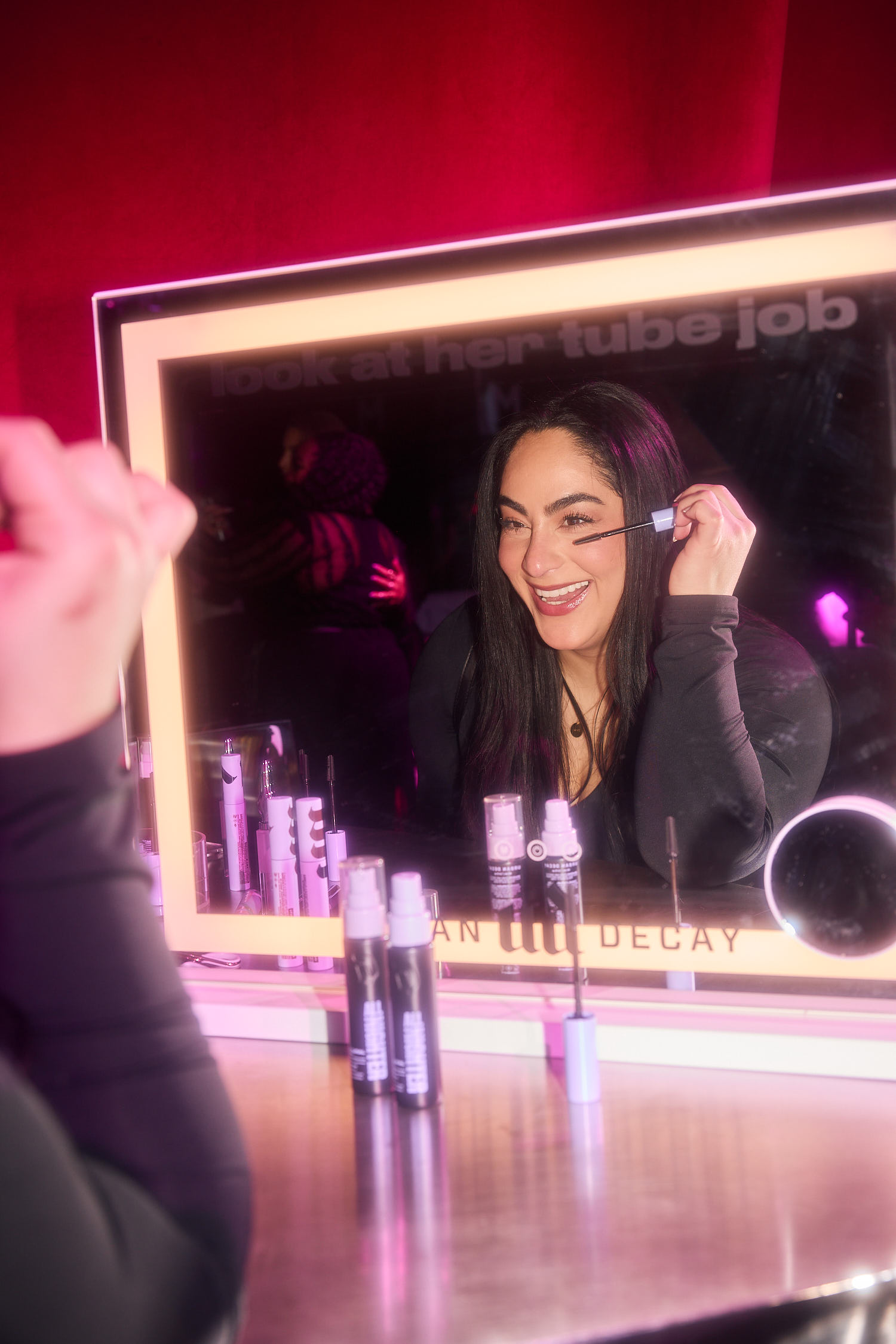 Woman applying makeup at a luminous vanity mirror, pink lighting, with makeup tubes on the counter in front of her