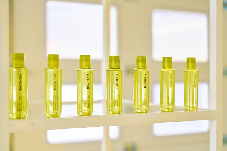 Row of small yellow cosmetic bottles lined on a white rack, backlit by bright windows in a spa-like display.