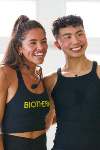 Two women in black athletic wear smiling together; one wears a Biotherm top and headset mic.