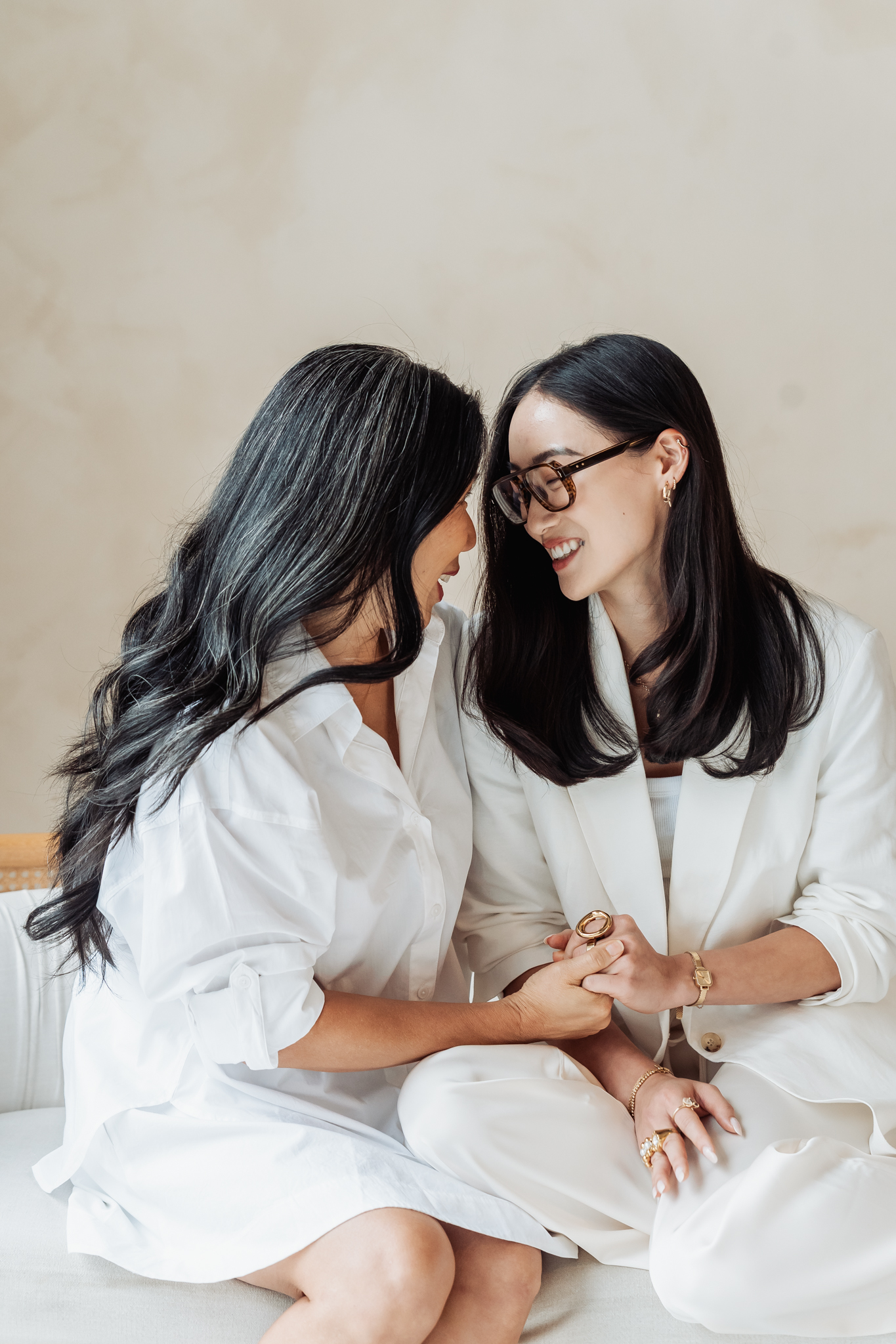 Two women in white outfits sit close together on a sofa, smiling at each other while exchanging a ring or jewelry piece.
