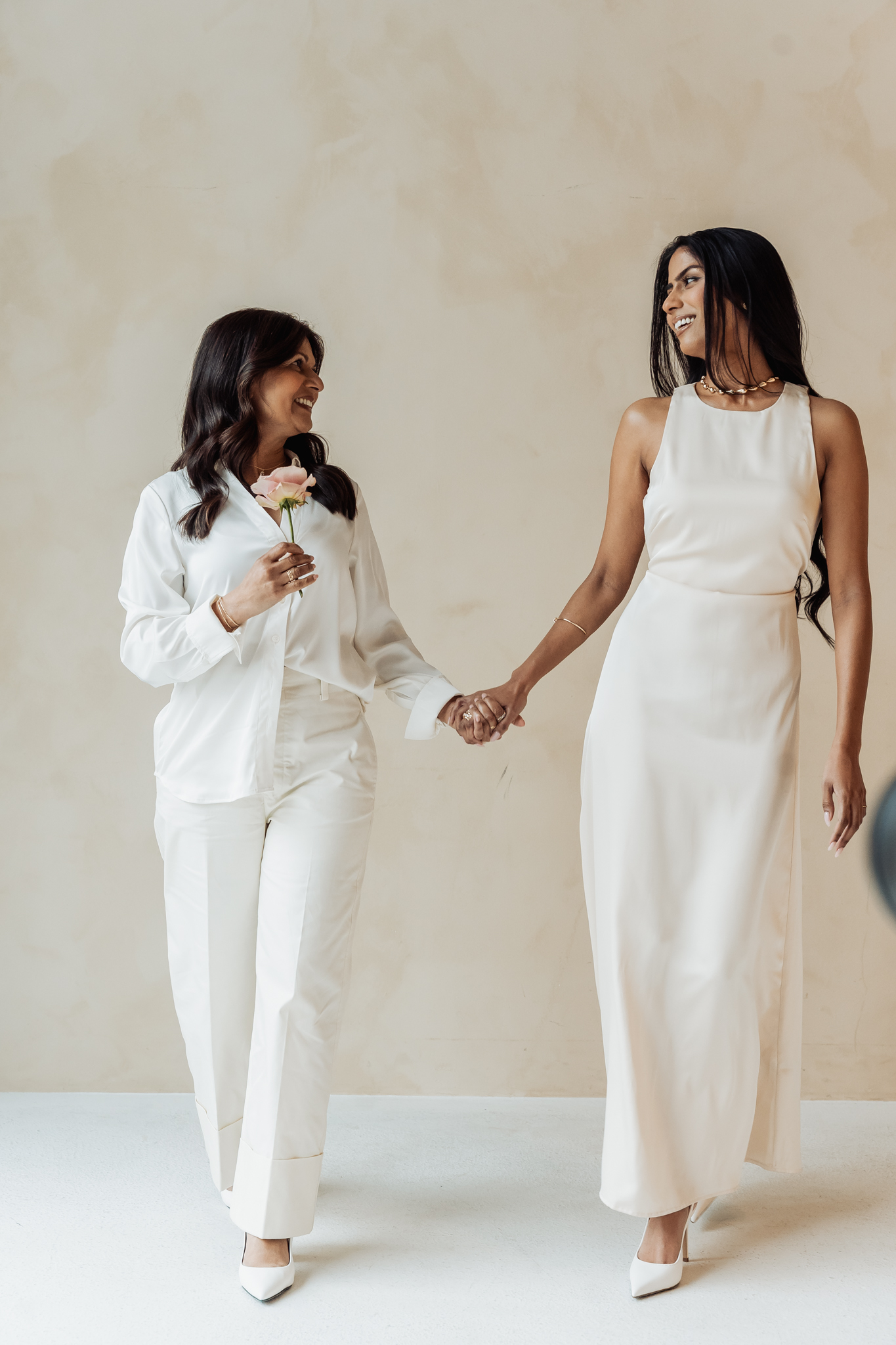Two women in white outfits smiling at each other while holding hands in a neutral room. One woman holds a pink flower.