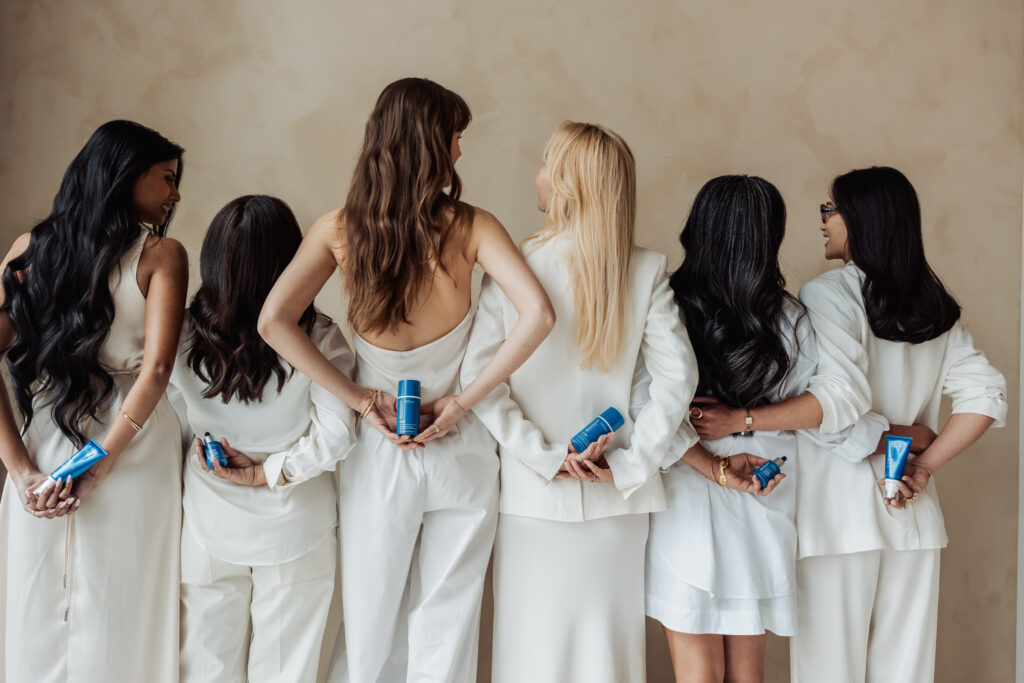 Group of women in white outfits standing with backs to camera, each holding blue bottles of hair products between them.