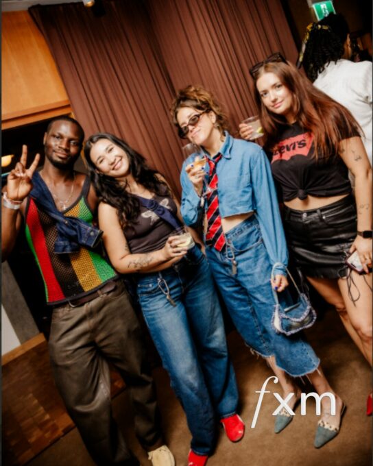 Four friends posing for a photo at a party, holding drinks and smiling at the camera, dressed in colorful casual outfits.