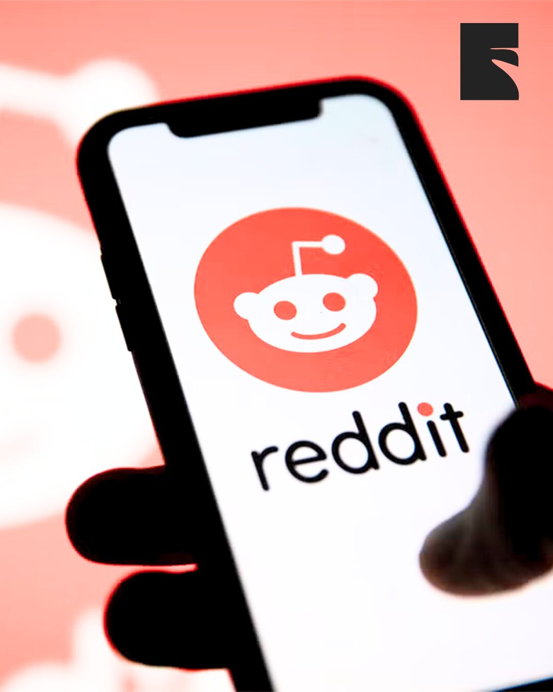 Smartphone displaying the Reddit app logo (orange circle with white Reddit alien) on screen, held in a hand.