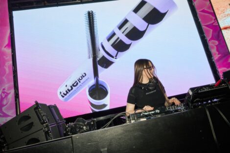 Female DJ in black shirt with headphones, mixing on a Pioneer DJ setup on a stage with a large screen showing makeup products behind her