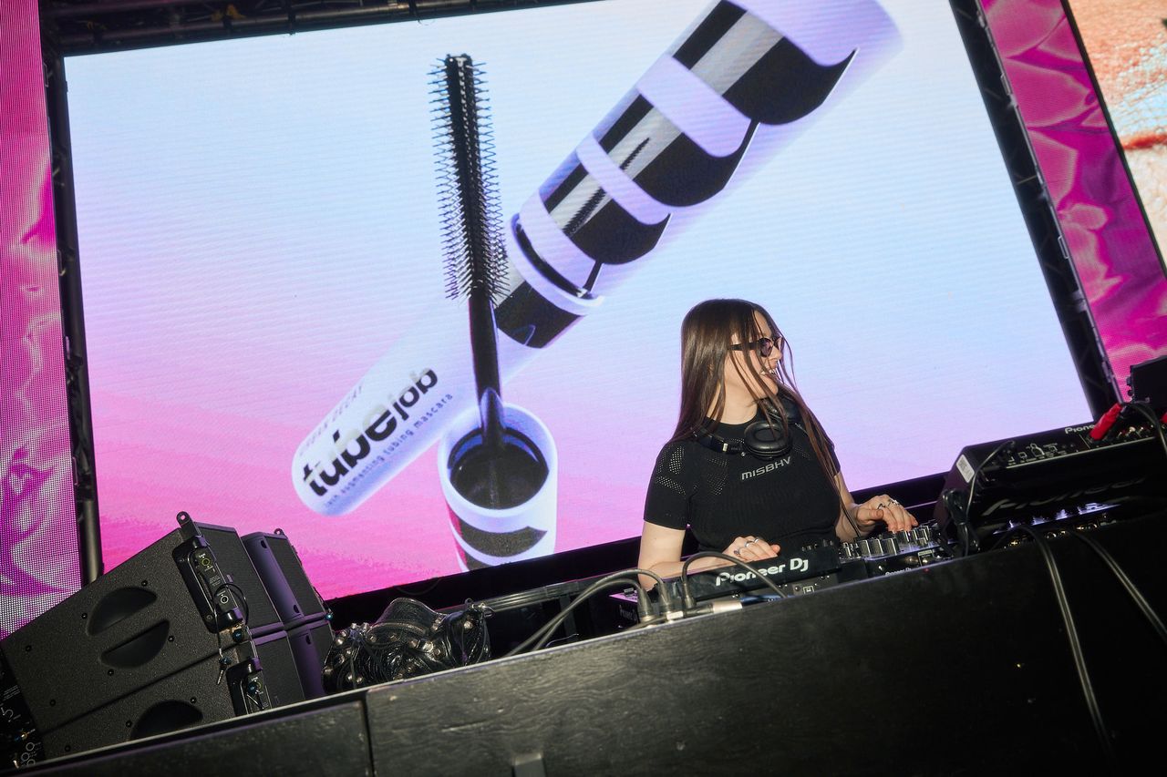 Female DJ in black shirt with headphones, mixing on a Pioneer DJ setup on a stage with a large screen showing makeup products behind her