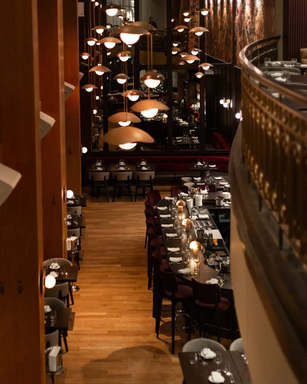 Elegant restaurant interior with long tables set for dining, warm pendant lights, and a wooden floor viewed from an upper balcony.